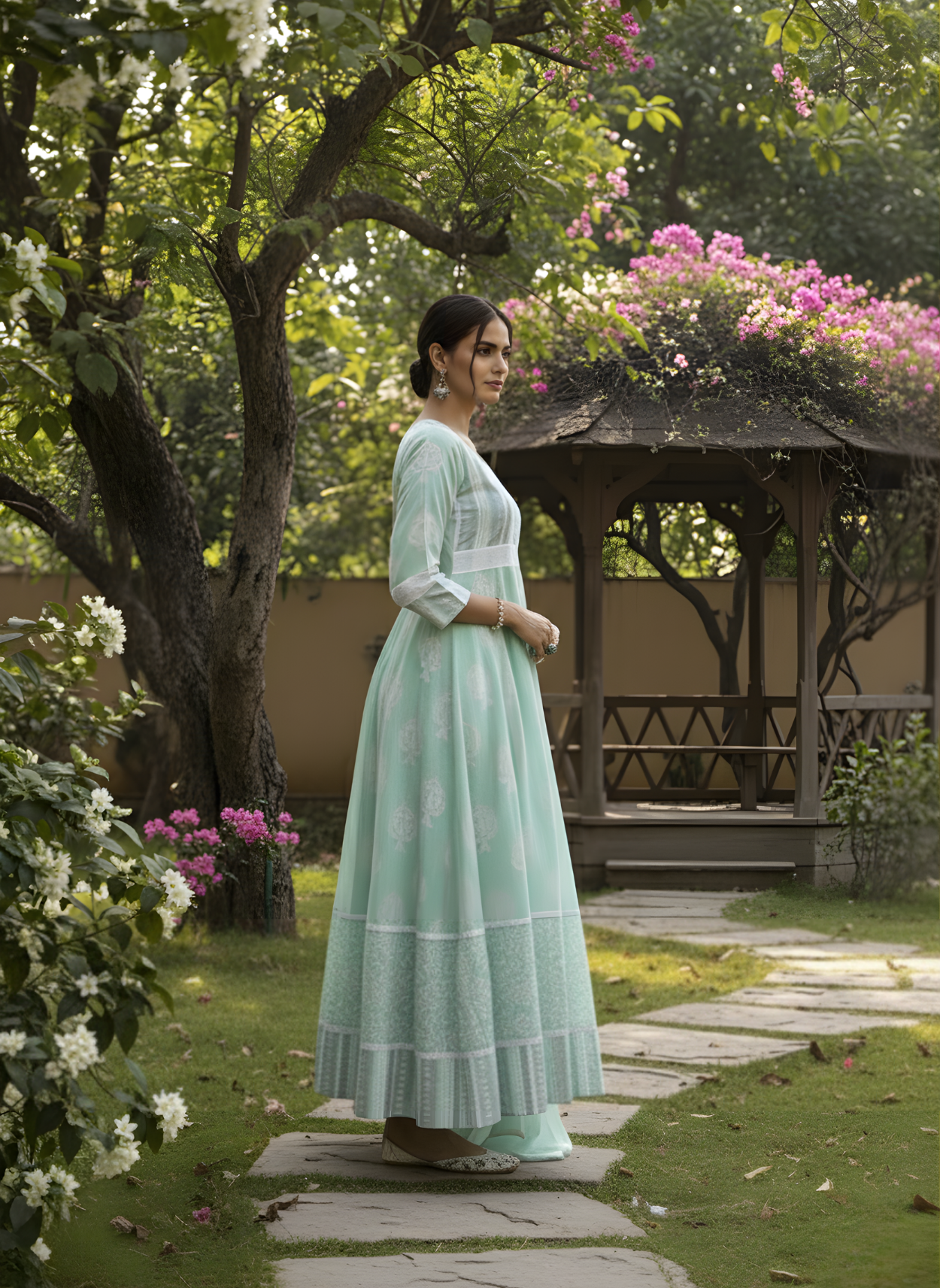 Woman in a light green dress standing in a garden with flowers and a gazebo.