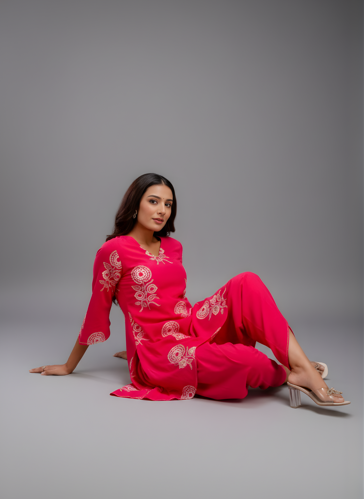 Woman in a bright pink outfit with white patterns sitting on a grey background