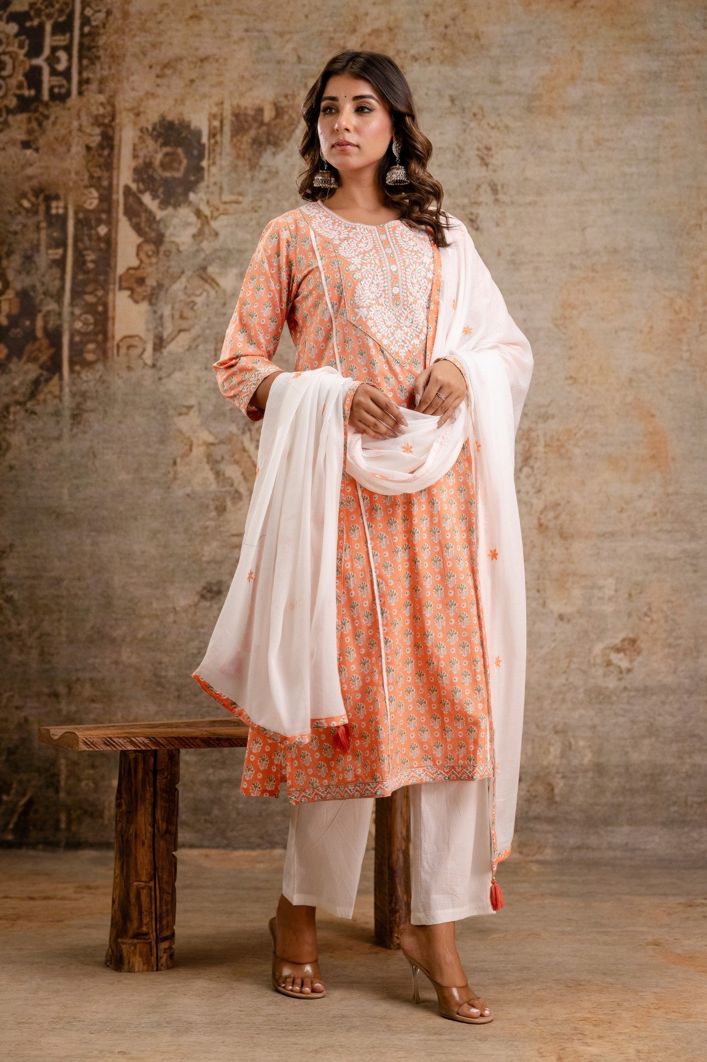 Woman in traditional outfit with orange and white patterned dress against a textured wall.