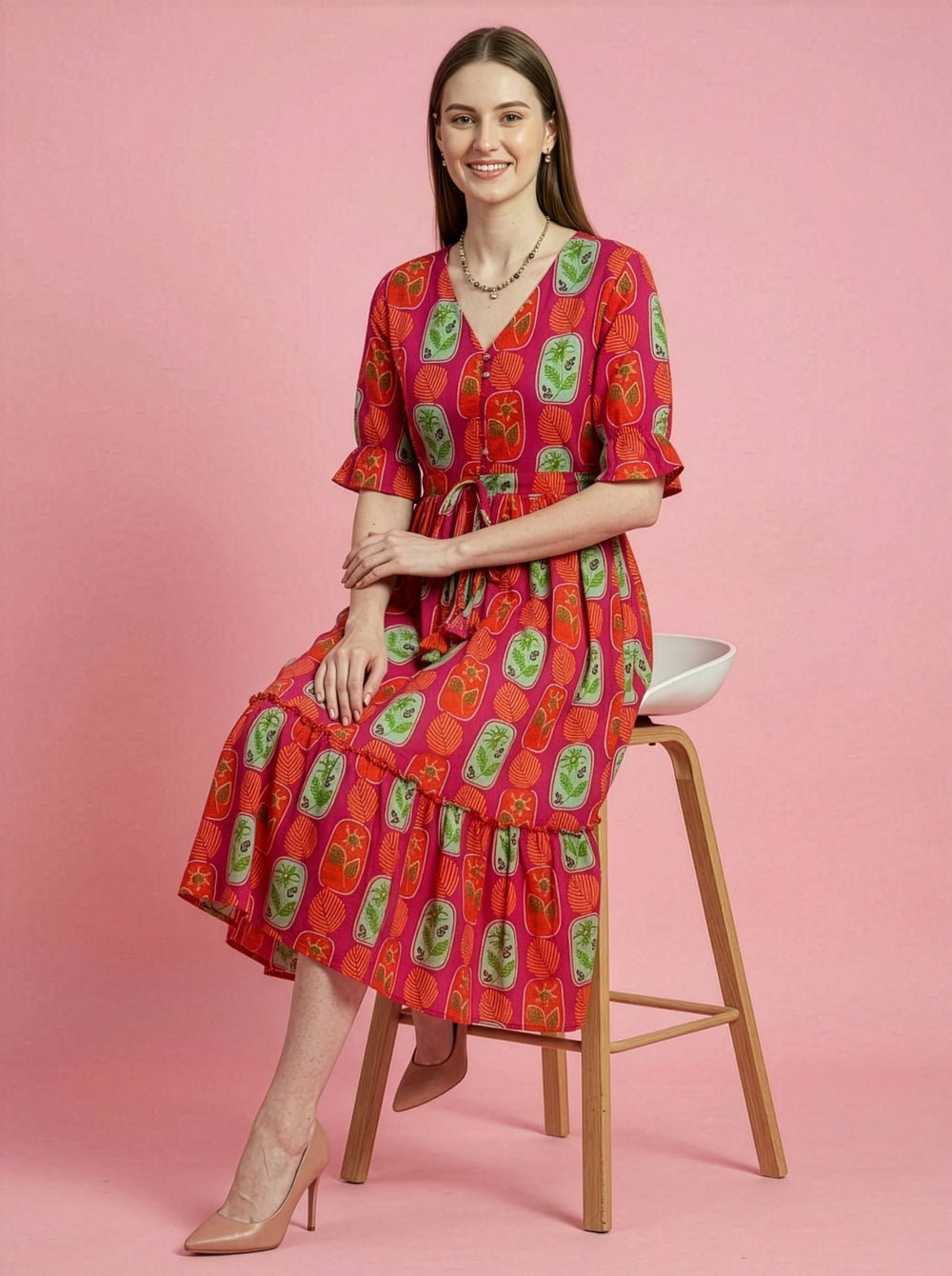 Woman wearing a red patterned dress sitting on a stool against a pink background
