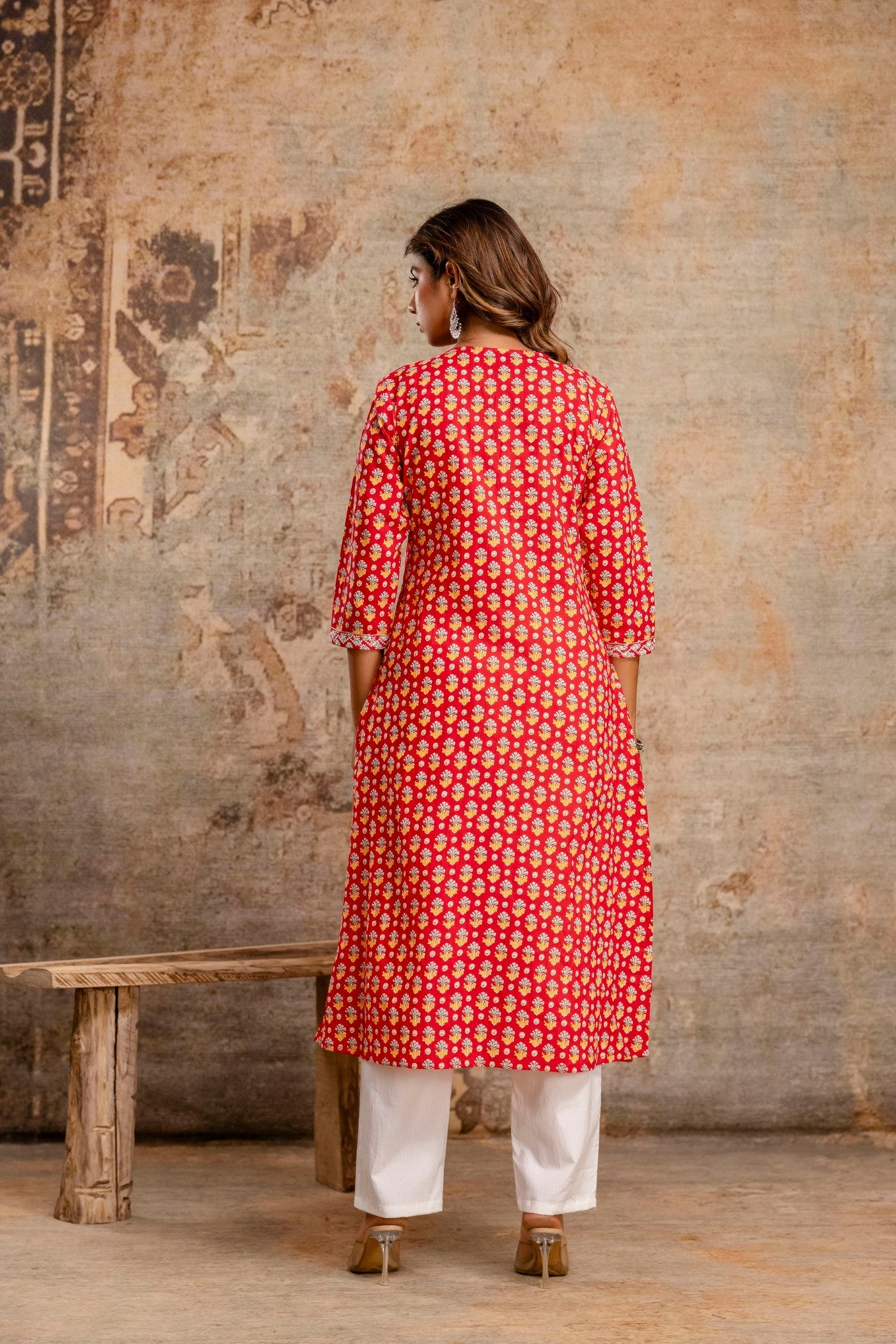 Woman wearing a red floral kurta with white pants standing against an aged wall.