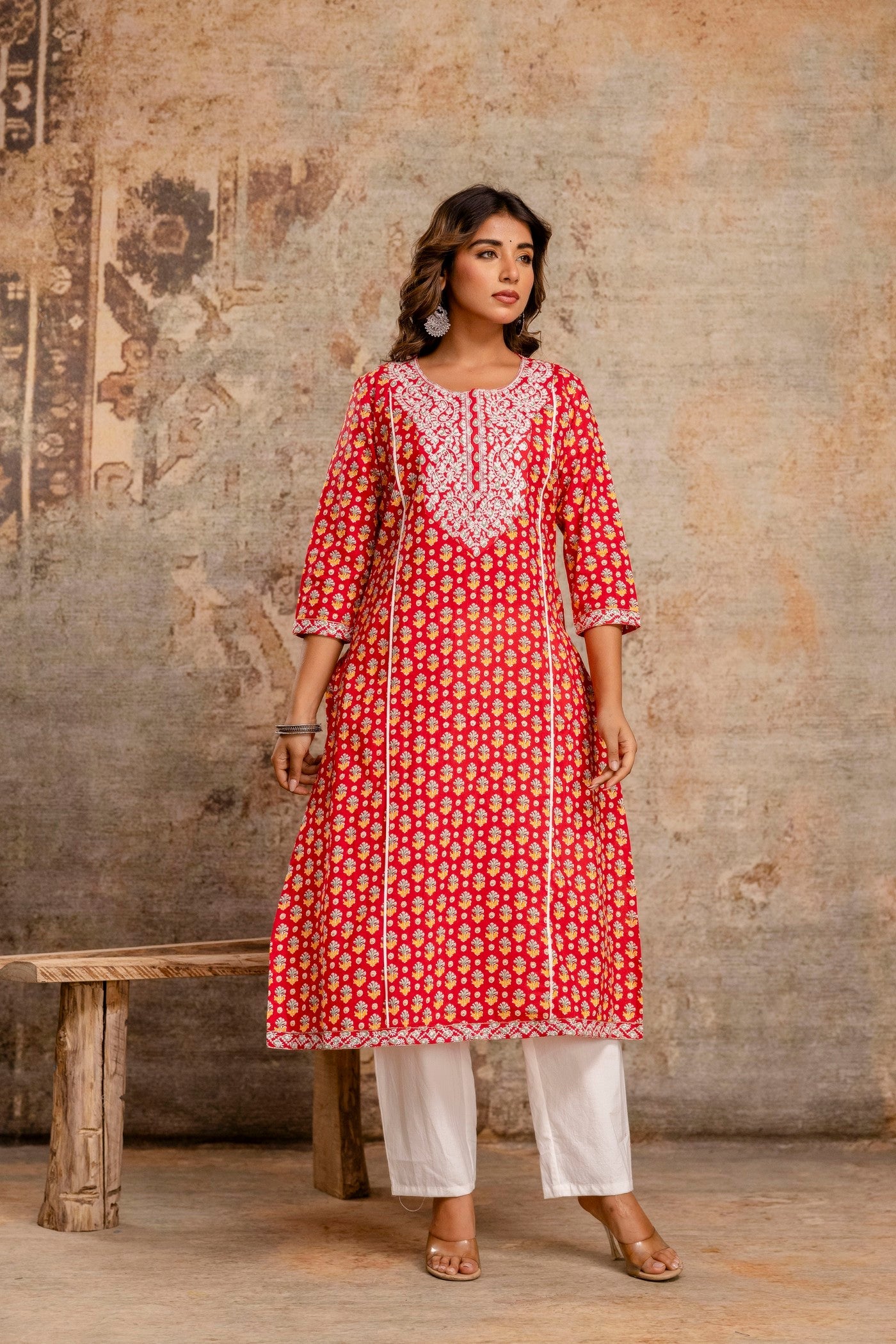 Woman wearing a red and white patterned kurta with white pants against a textured wall.