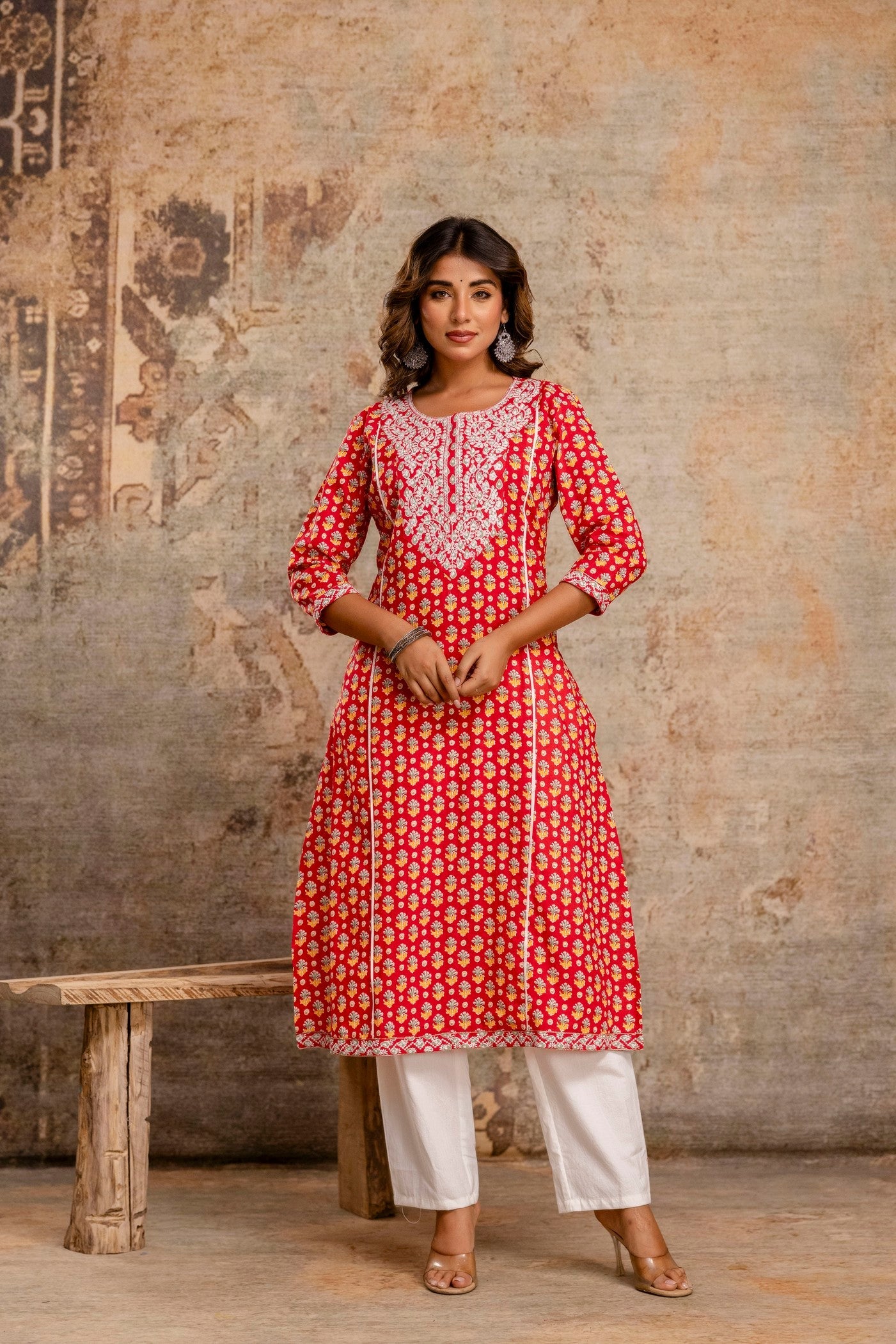 Woman wearing a red and white patterned kurta with white pants against a textured wall.