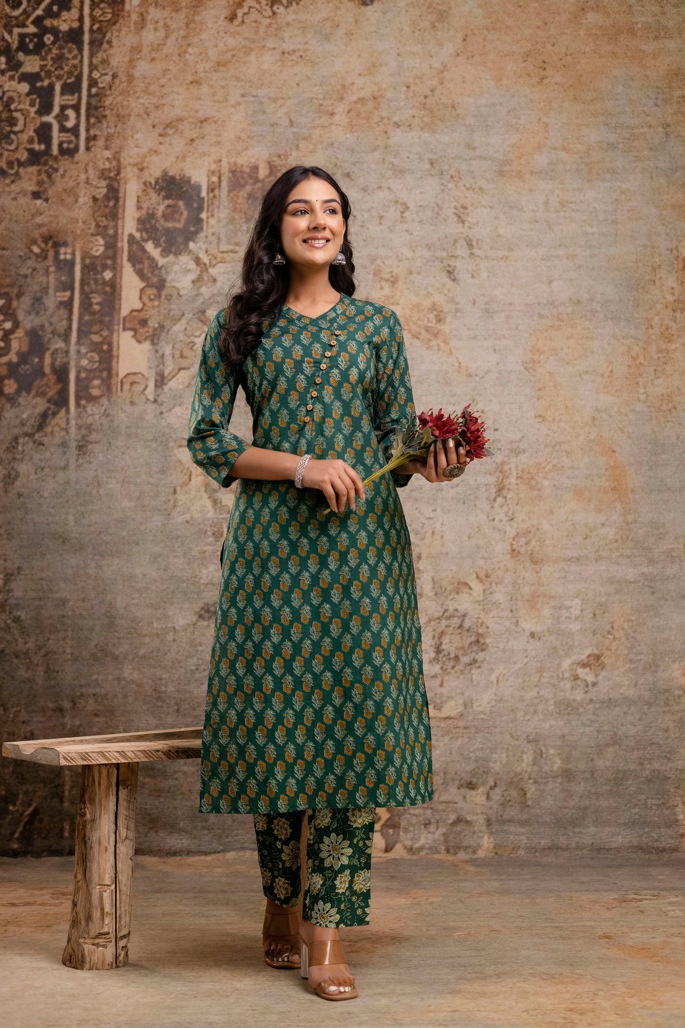 Woman in a green patterned outfit holding flowers against a textured wall.